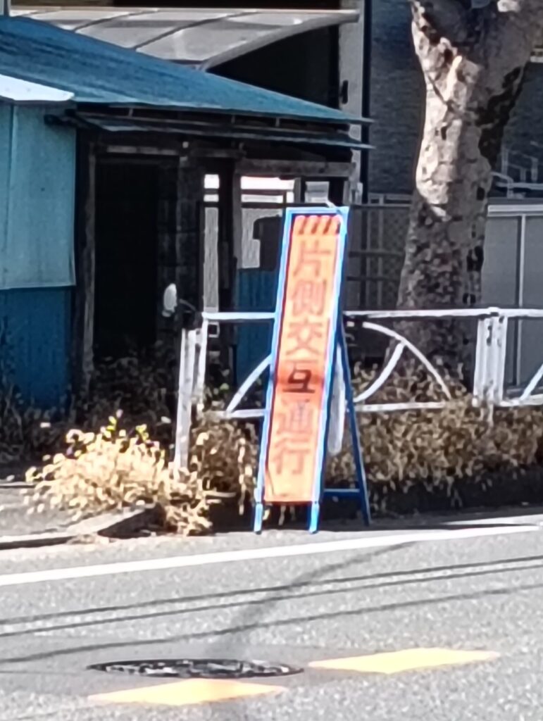 工事現場手前の道路に設置された『片側交互通行』の案内看板。通行車両へ注意を促す、オレンジ色と黒の警戒標識の風景。